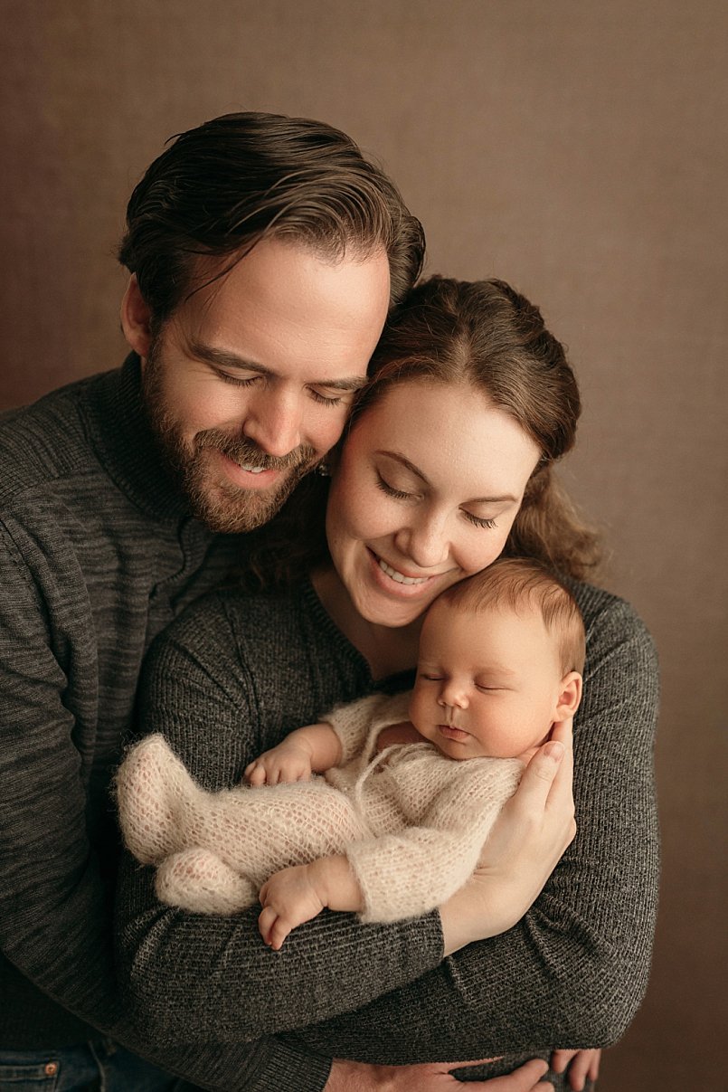 A tender studio portrait by a Portland newborn photographer featuring a mother and father smiling at their sleeping baby dressed in a soft, cream-colored knit outfit.