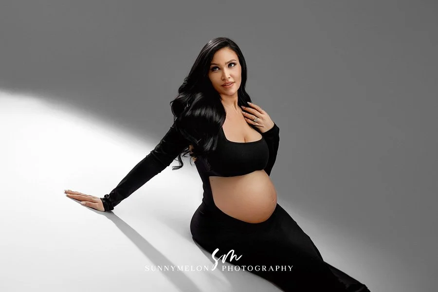 A side-profile studio portrait of a pregnant woman wearing a sleek black long-sleeve bodysuit, reclining against a minimalist grey background with dramatic lighting.