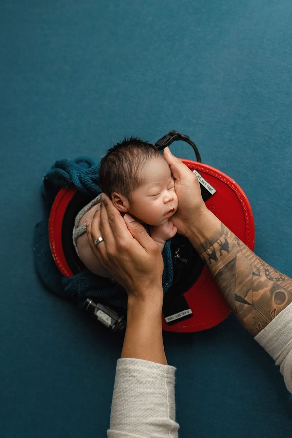 A professional overhead studio newborn portrait by SunnyMelon Photography featuring a sleeping infant posed inside a red and black firefighter helmet. The baby is resting peacefully on a dark blue textured backdrop, with a pair of tattooed hands gent