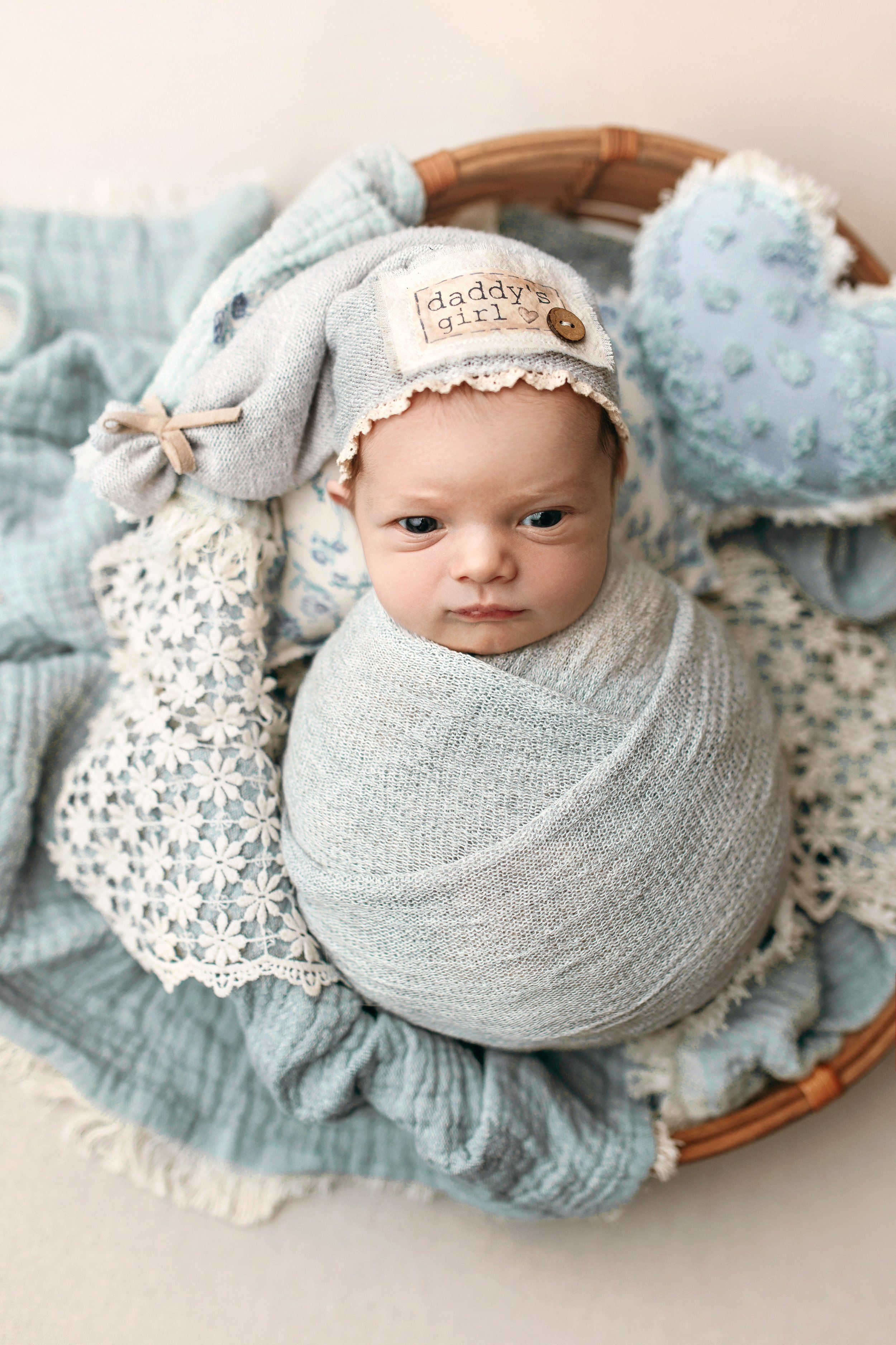 A professional studio newborn portrait of a baby girl nestled in a rustic wicker basket. The infant is tightly swaddled in a soft light blue textured wrap and wears a matching long-tail sleepy hat with a "daddy's girl" patch. The basket is layered wi