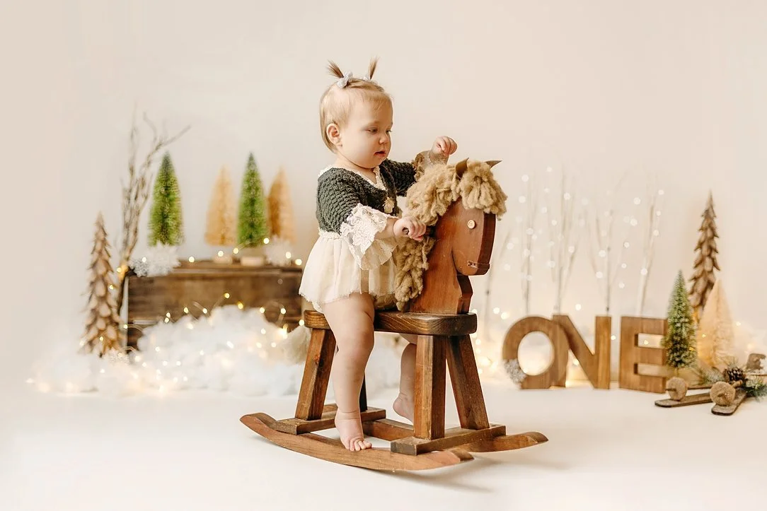 This whimsical milestone portrait features a young toddler celebrating her first birthday in a beautifully styled "Winter Wonderland" set. The child is captured sitting on a rustic vintage rocking horse, dressed in a delicate dark green knit and crea