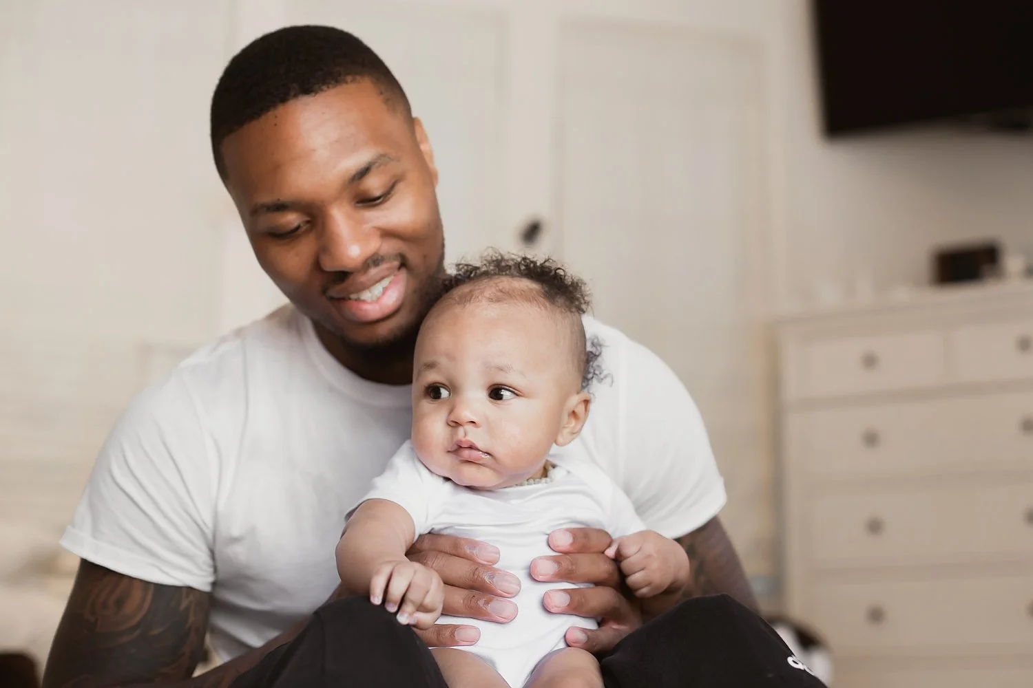 NBA star Damian Lillard smiling while holding his infant son, Dame Jr., during a lifestyle newborn photography session in a bright, modern home setting.