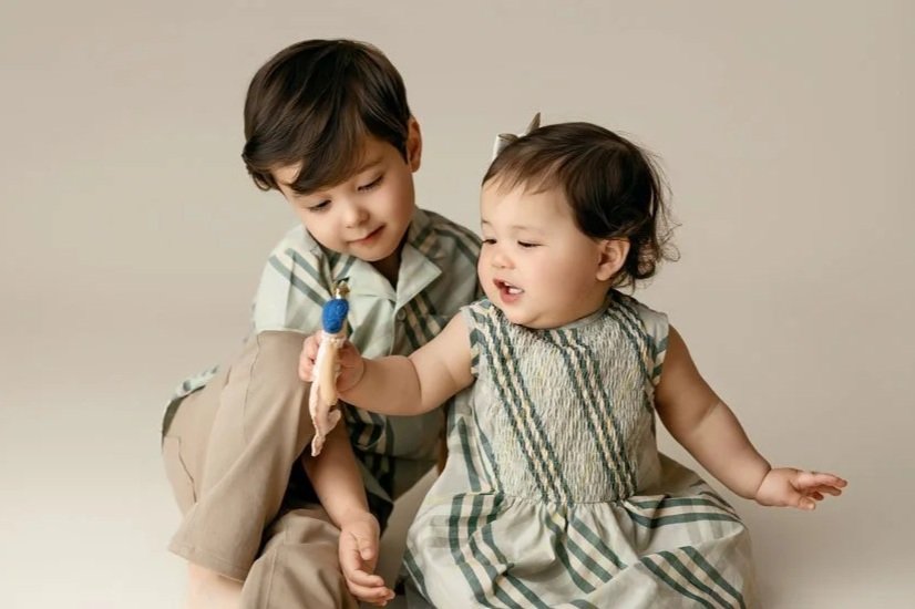 A studio portrait of a young boy and his younger sister, both wearing coordinated plaid outfits, sitting against a warm, neutral painted background. Baby girl holds a small toy, smiling at it.