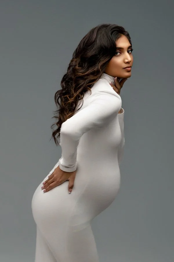 Profile portrait of a pregnant woman with long wavy dark hair, wearing a sleek white long-sleeve turtleneck maternity dress, posing against a neutral gray studio background.
