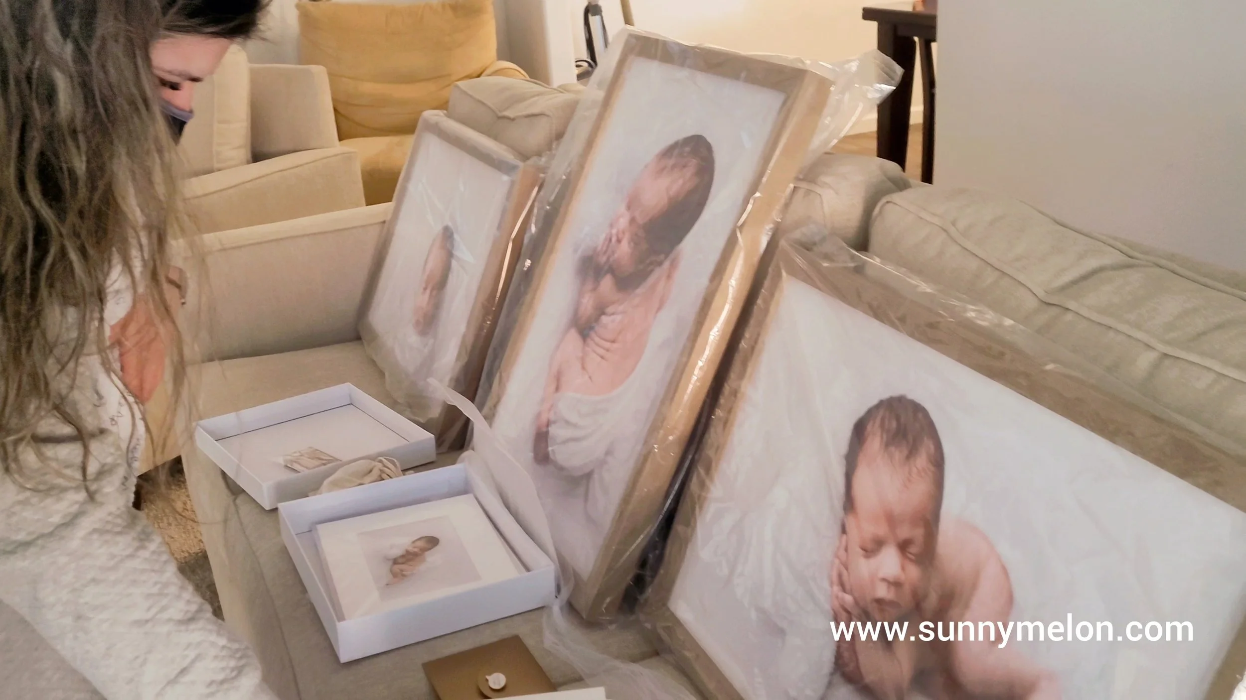 A collection of high-end, professionally framed newborn portraits still in their protective packaging, resting on a sofa. A woman leans in to inspect the fine details of the prints, showcasing the scale and quality of custom-ordered gallery wall art.