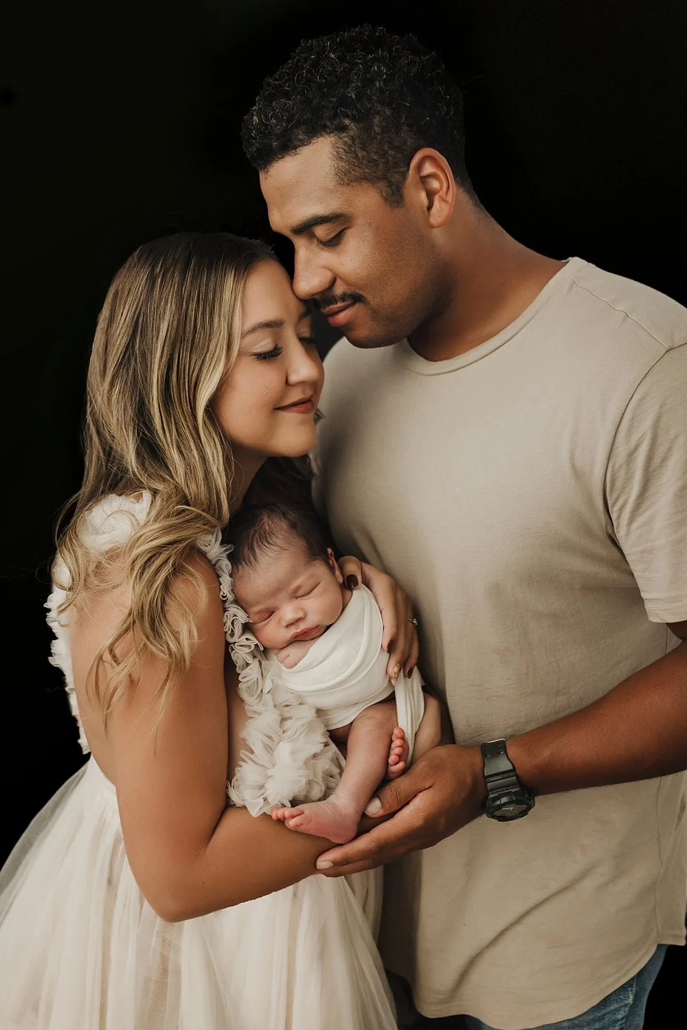A professional newborn family photograph featuring a mother and father in close proximity. The mother, wearing a white ruffled dress, cradles the infant who is snugly swaddled in a white wrap. The father, wearing a tan t-shirt and a black watch, lean