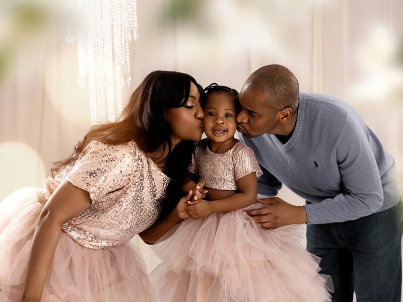 A family of three, including a young girl and her parents, sharing a kiss and a hug while dressed in pastel pink and blue clothes inside a warmly lit room.