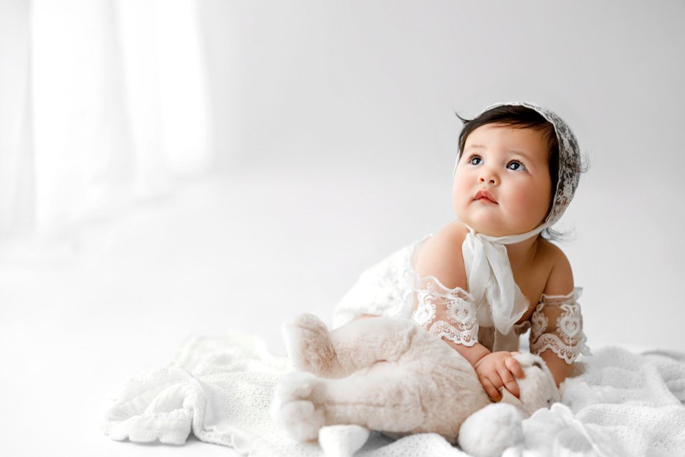 A professional high-key studio portrait of a baby girl wearing a delicate lace bonnet and matching outfit, sitting on a soft white blanket with a plush teddy bear.