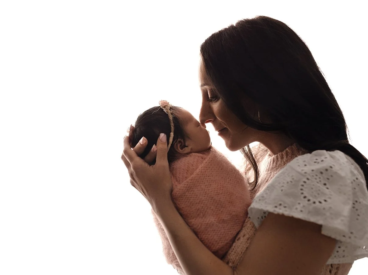 A side-profile photograph of a mother holding her newborn baby, who is wrapped in a soft pink swaddle, against a solid white studio background.