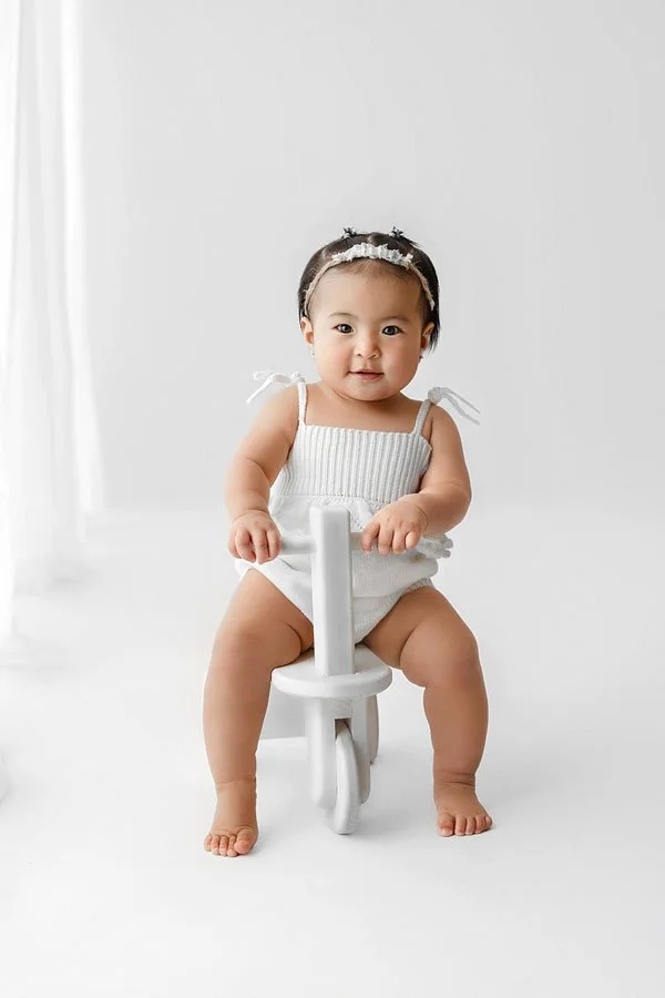 A baby girl in a white knit romper and headband sitting on a white wooden toy tricycle in a bright, minimalist photography studio.