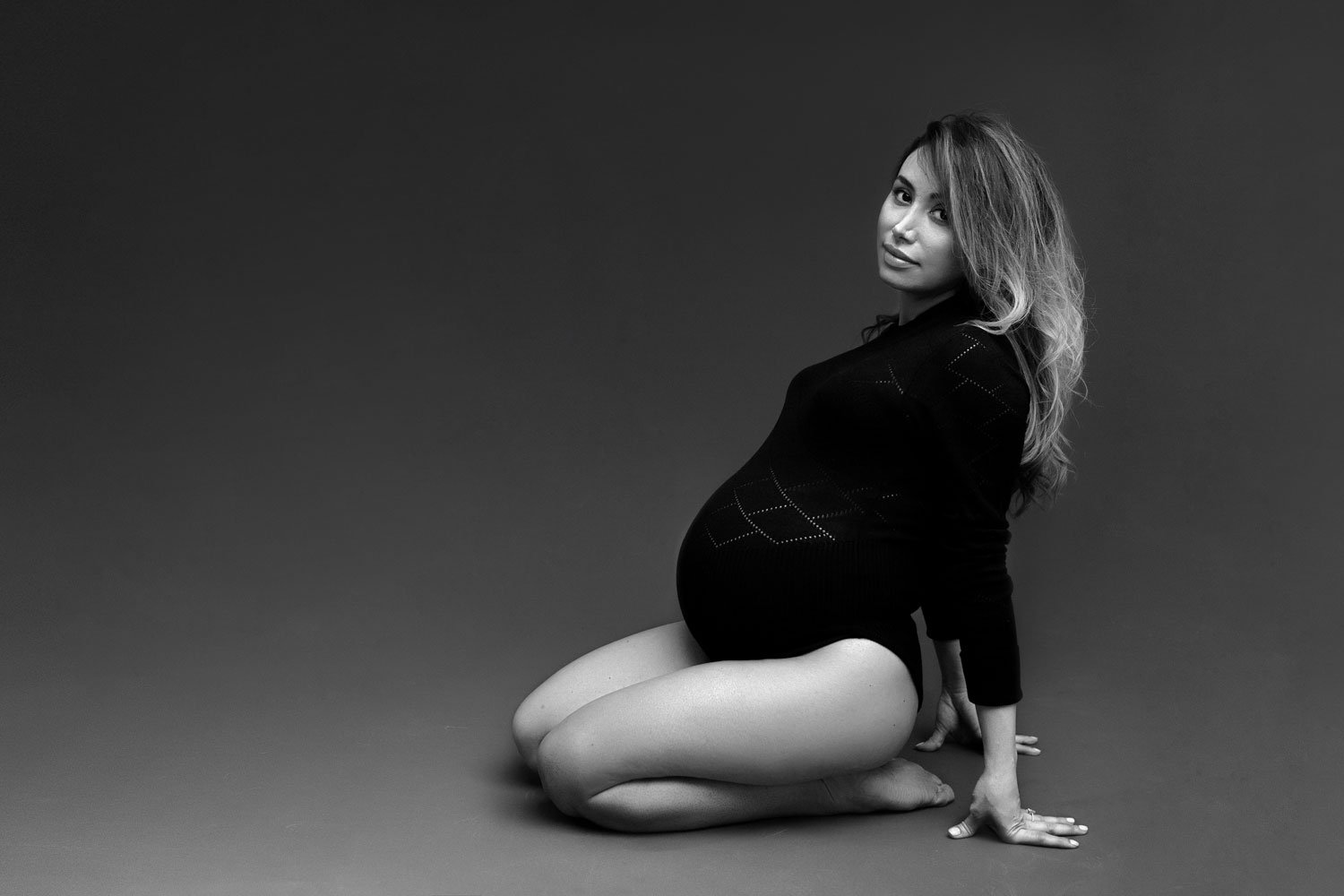 Black and white studio maternity portrait of a pregnant woman in a black bodysuit kneeling against a dark backdrop in West Linn, Oregon, showcasing her contoured silhouette.