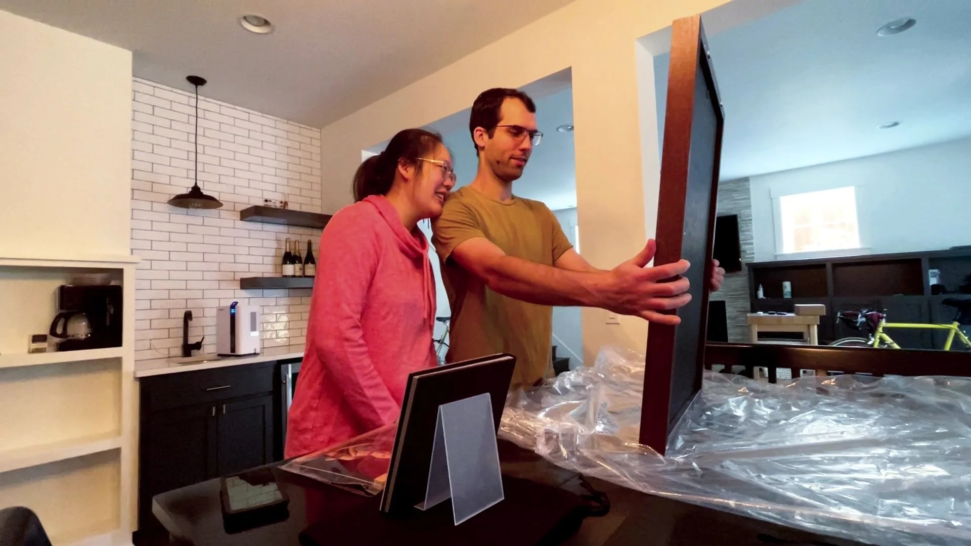 A candid shot of a happy couple in their kitchen excitedly unboxing a large, high-quality framed family portrait. The image highlights the premium physical products and personalized gallery reveal service provided by a professional Portland family ph