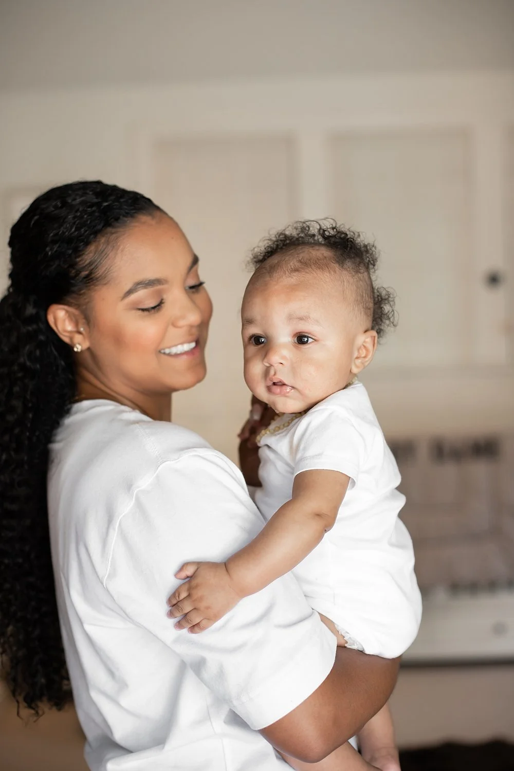 A heartwarming lifestyle portrait featuring Kayla Lillard looking affectionately at her infant son, Dame Jr. Kayla has long, dark curly hair and is wearing a white long-sleeve shirt. The baby is wearing a white onesie and looking off-camera. The back