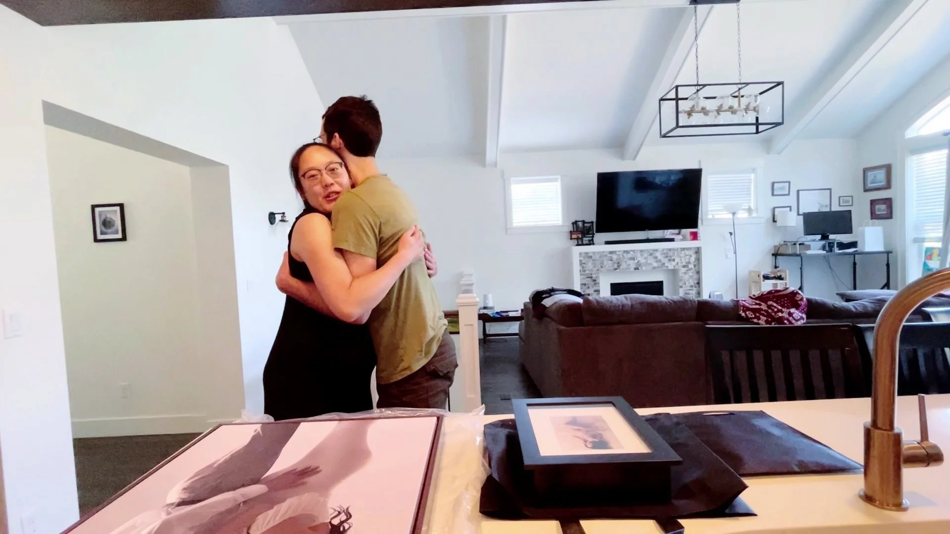 A poignant wide shot of a couple sharing a heartfelt hug in their bright, modern living room. In the foreground, newly delivered professional wall art and framed portraits rest on a counter, still in their packaging. The scene perfectly captures the 