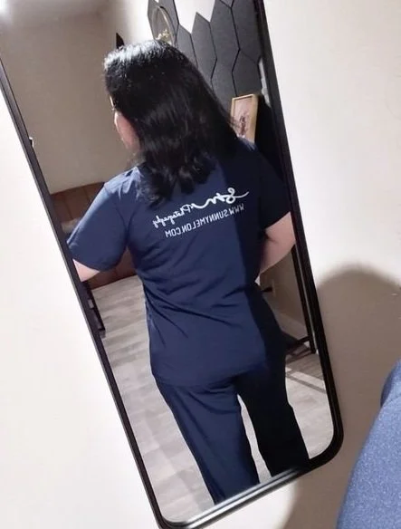 A mirror selfie from behind showing a person with dark hair wearing a navy blue t-shirt printed with the white script logo and website URL for SunnyMelon Photography.