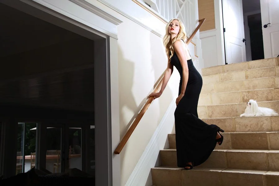 A blonde woman in a black evening gown posing on a grand stone staircase alongside a white Maltese dog in a luxury home setting.