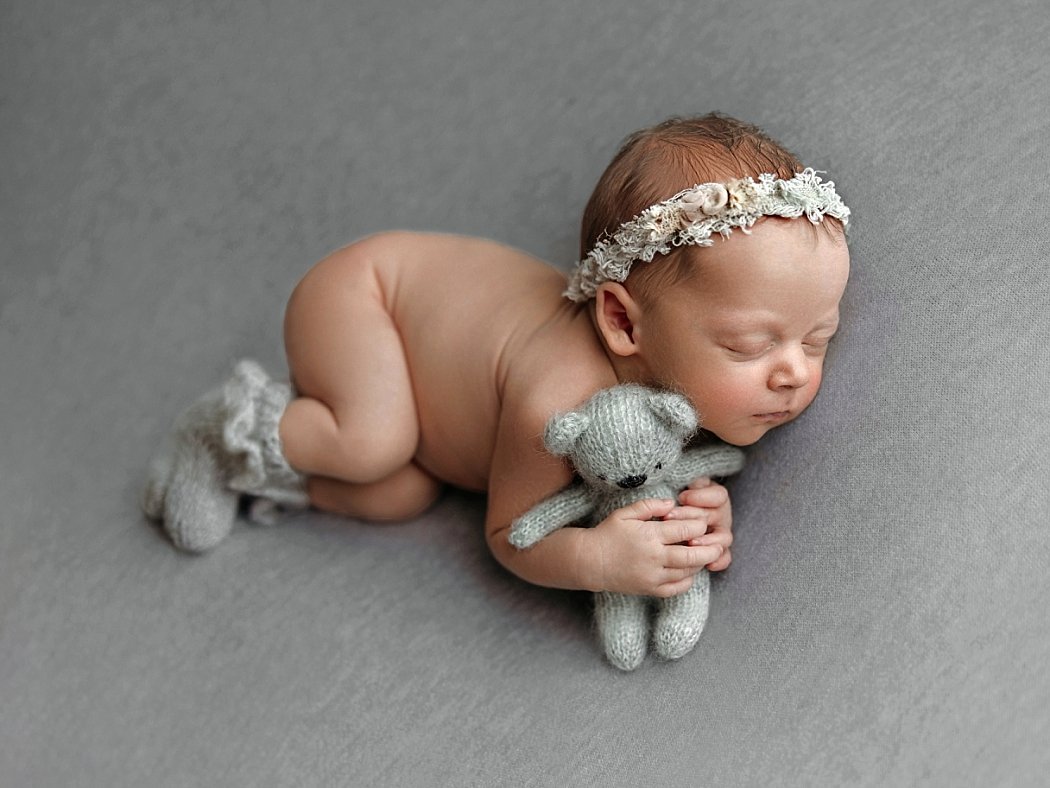 A sleeping newborn baby posed on their stomach against a solid gray backdrop, wearing a delicate floral lace headband and tiny knit socks while holding a small gray knit teddy bear.