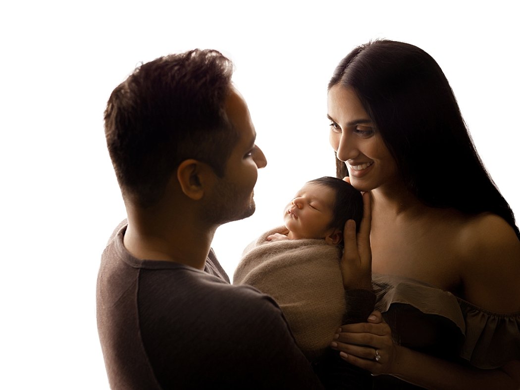 A heartwarming family portrait using high-contrast backlighting. The father, seen in profile from the left, and the mother, with long dark hair on the right, look affectionately at their infant. The newborn is peacefully asleep and wrapped in a soft,