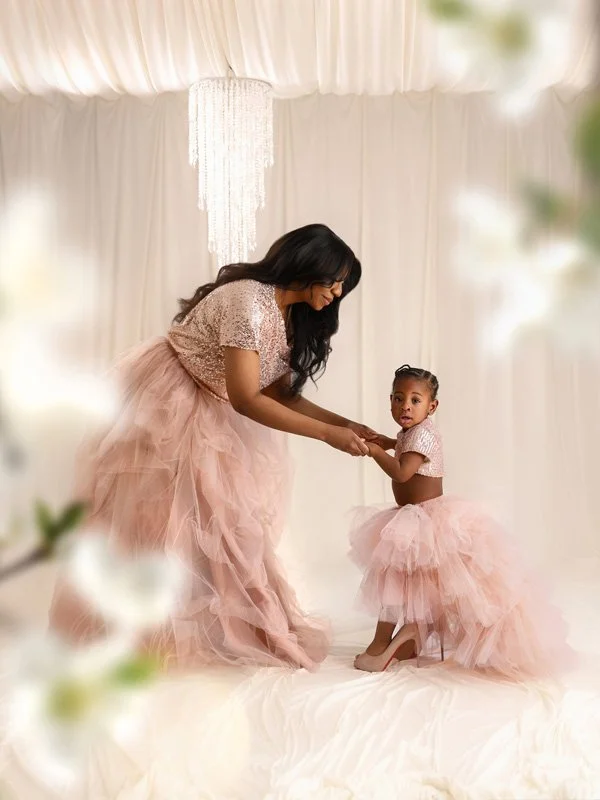 A mother in a rose gold sequin top and pink tulle skirt holding her young daughter's hands as the toddler balances in oversized high-heeled shoes in a bright, ethereal studio.