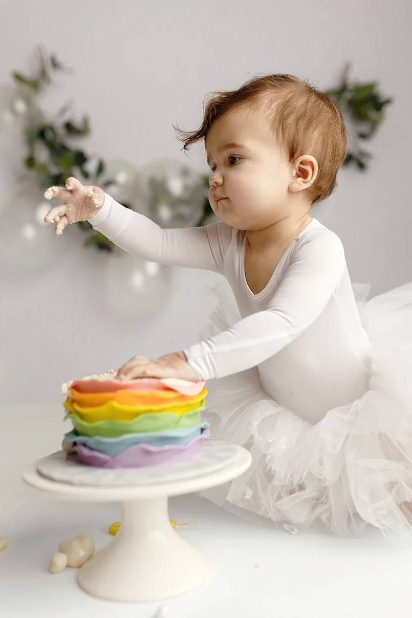 This joyful first birthday portrait captures a classic "cake smash" moment in a bright, airy studio setting. The toddler is dressed in a clean white long-sleeved leotard and a fluffy white tulle tutu, reaching playfully toward a vibrant, multi-colore