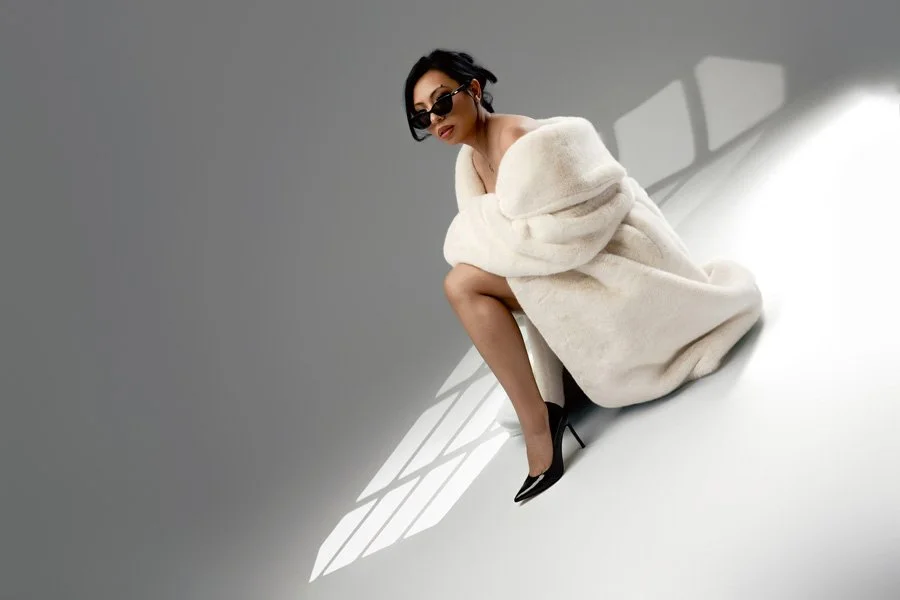 A full-length editorial fashion portrait of a woman crouched in a minimalist studio, wearing a large white faux-fur coat and sunglasses, with dramatic spotlight patterns on the white floor and a grey background.
