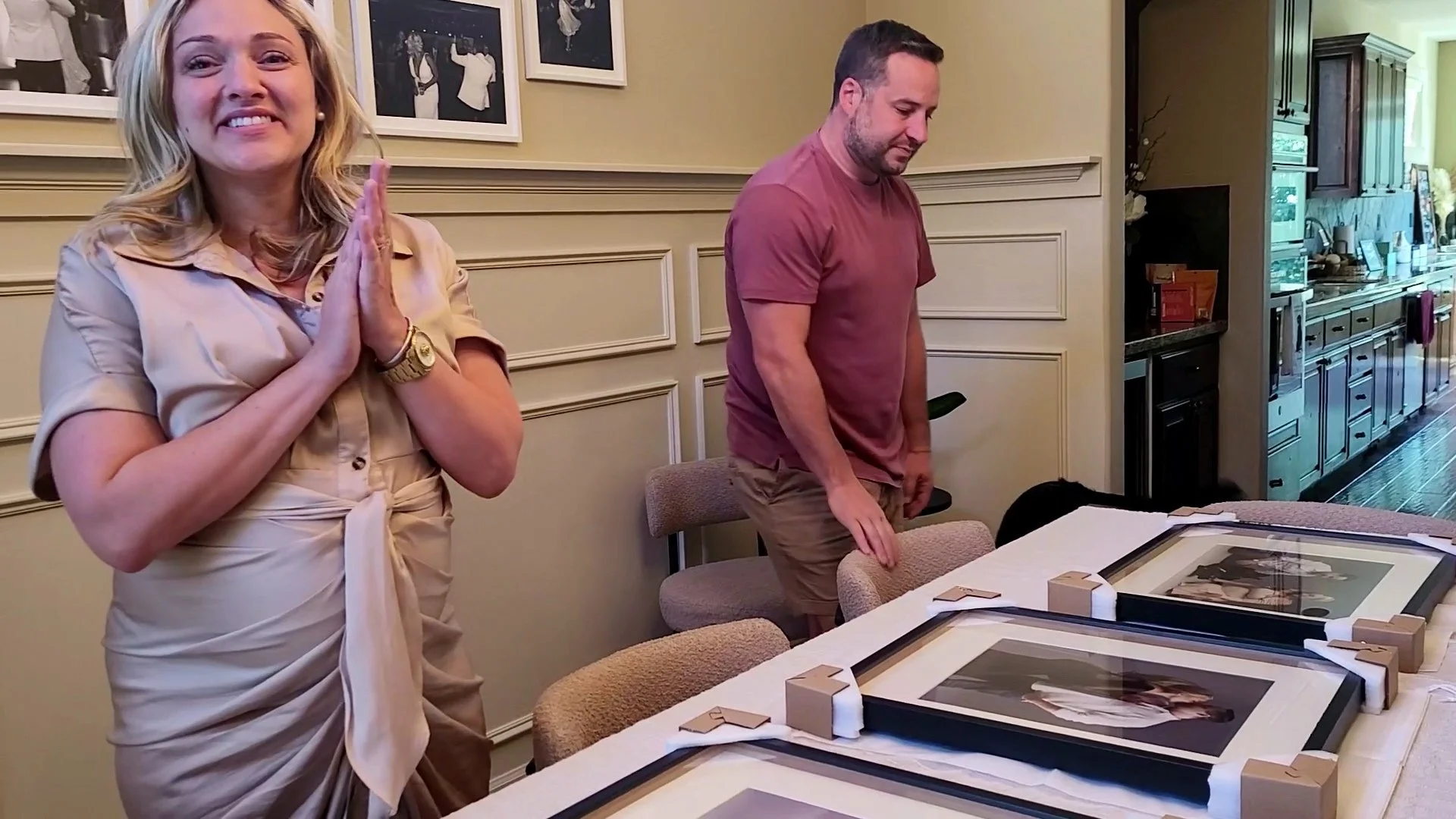 A woman with a joyful expression and hands clasped in gratitude stands next to a man reviewing several large, black-framed professional portraits laid out on a white table.