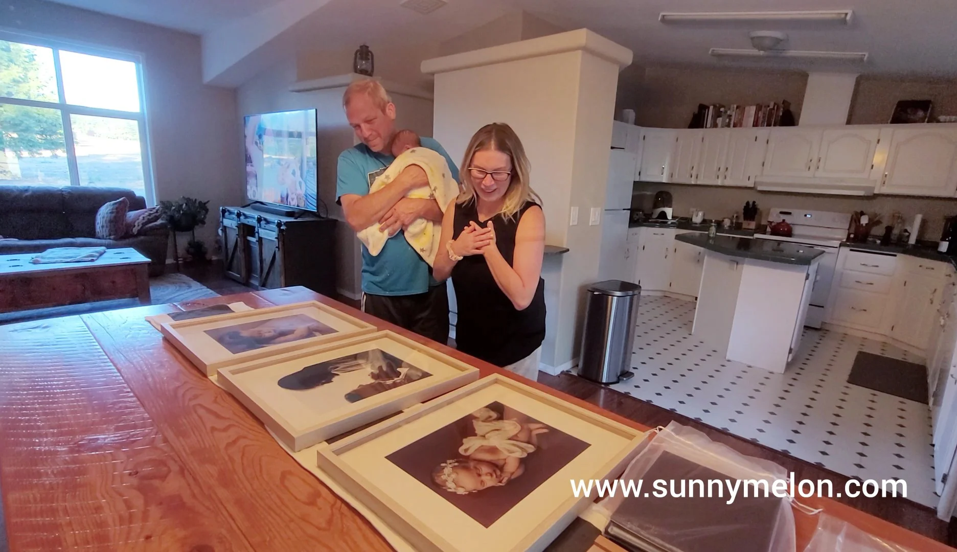 This candid shot captures the poignant moment of an in-home photography reveal, showing a mother's visible emotion as she sees her newborn’s portraits for the first time. The father holds the sleeping infant while they admire three custom-framed fine