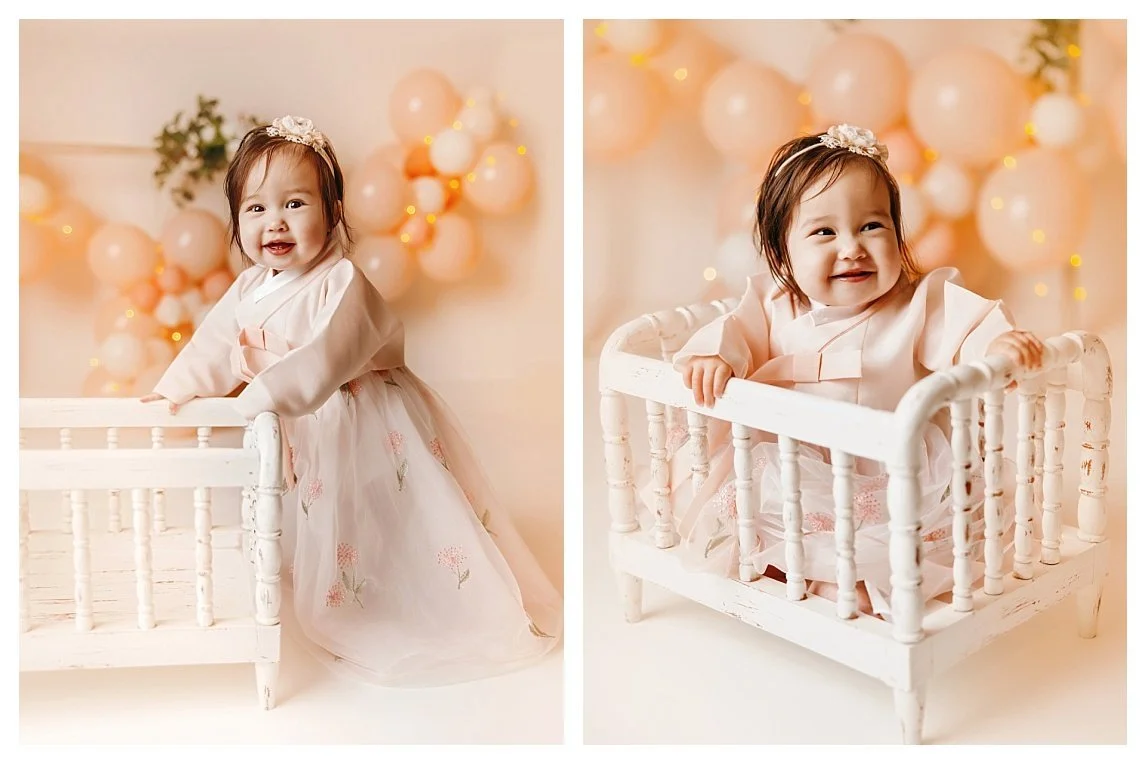 A two-image collage of a baby girl in a pink and white traditional Korean Hanbok; she stands beside a vintage white wooden crib prop on the left and sits inside it on the right against a cream backdrop.