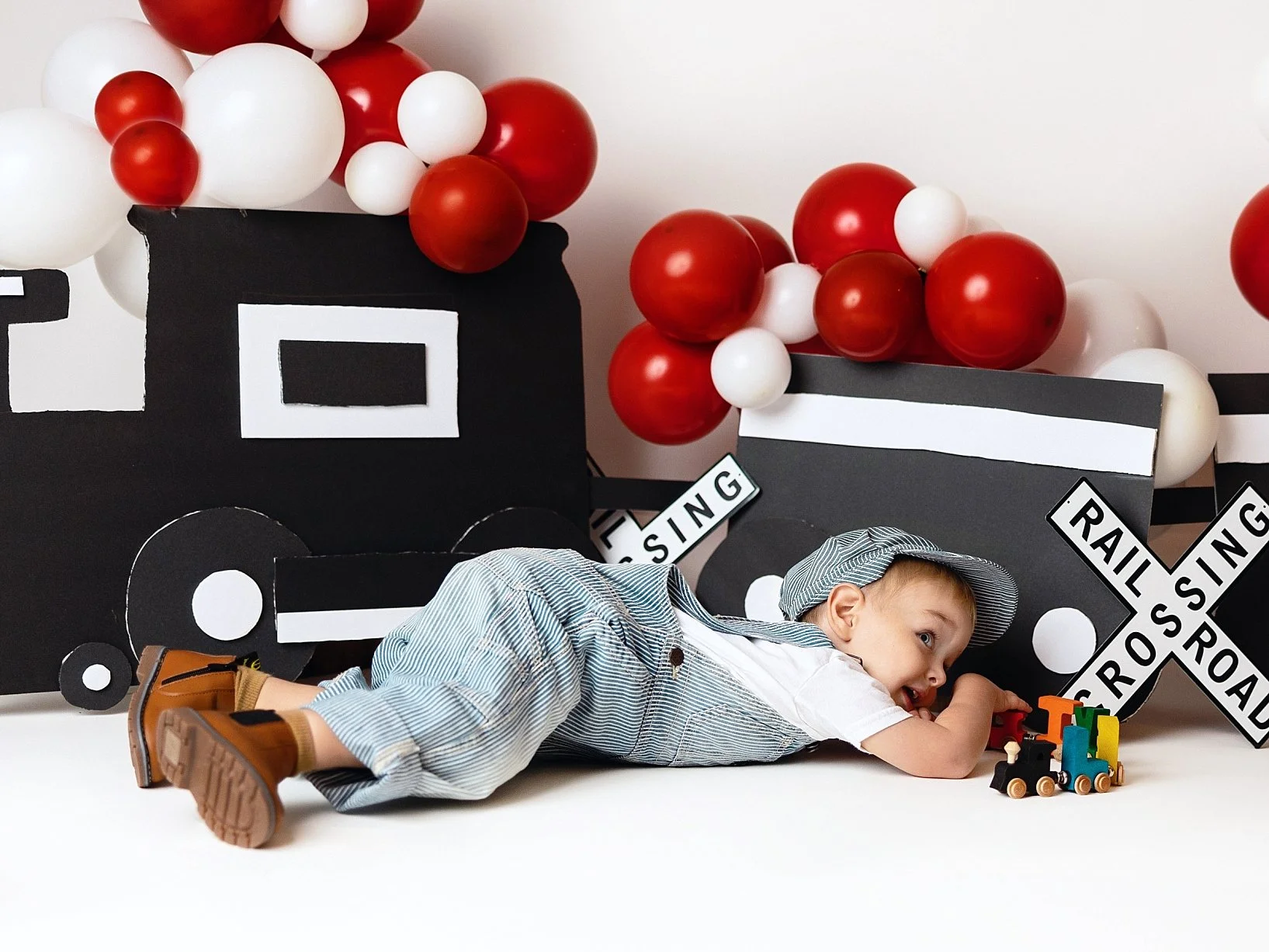 This imaginative milestone portrait features a young boy immersed in a playful "all aboard" train-themed photoshoot. The toddler is captured laying on his stomach, dressed in classic hickory stripe overalls and a matching conductor's cap, while focus