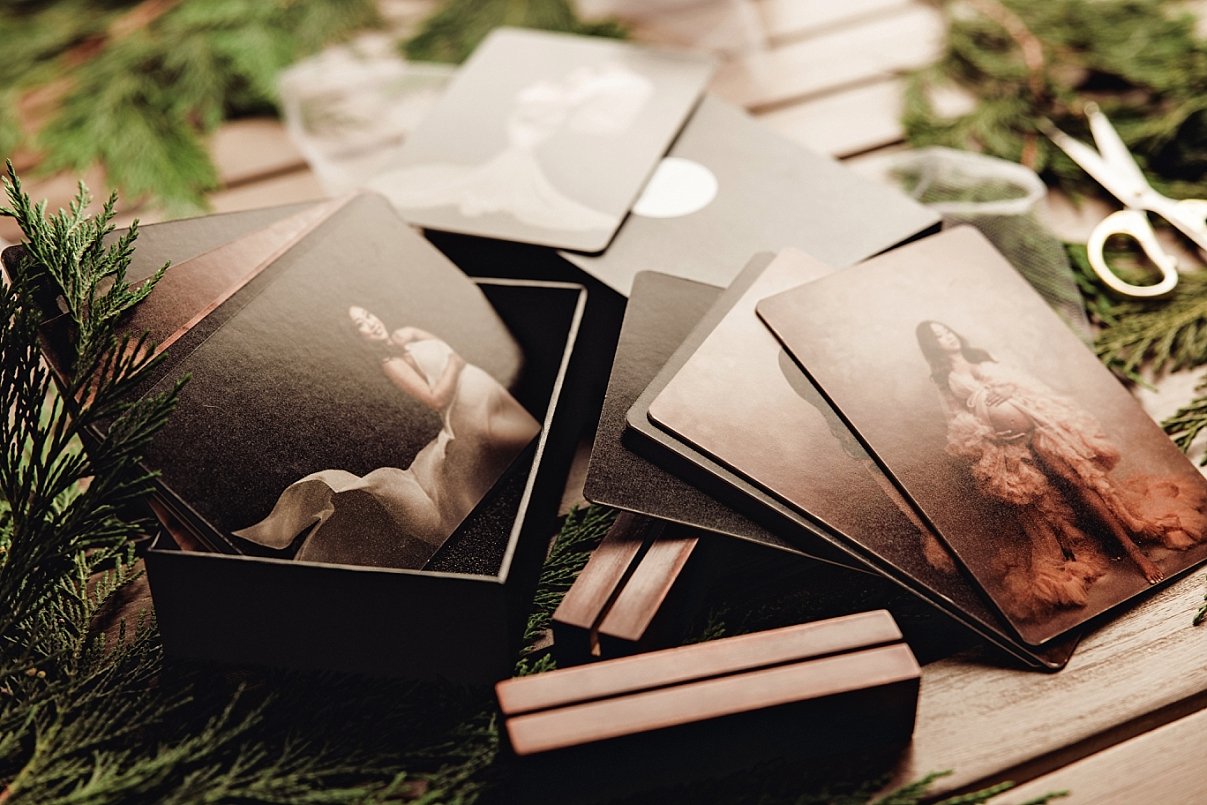 A collection of professional fine art maternity prints featuring a woman in elegant gowns, displayed in a luxury black folio box on a wooden surface with greenery and gold scissors.
