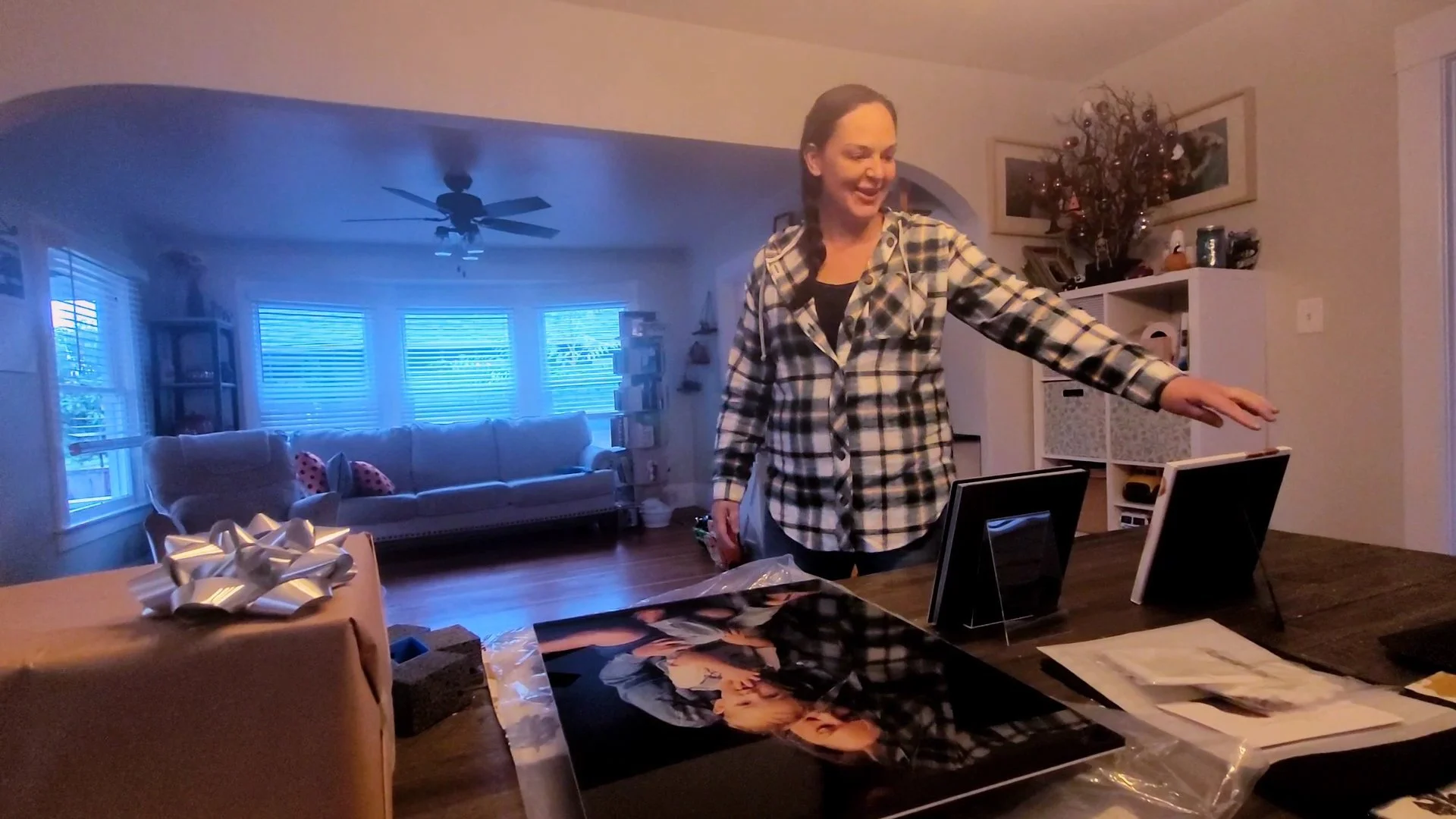 A smiling woman in a plaid shirt presents high-quality framed family portraits in her living room. The scene showcases the final physical products and personalized gallery reveal experience provided by a Portland family photographer.
