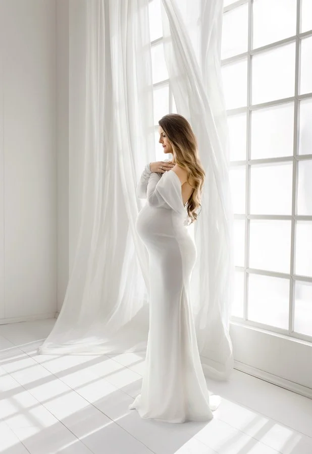 A high-end studio maternity portrait of a woman with long wavy hair, wearing a form-fitting white off-the-shoulder gown, standing in profile in front of a large window with sheer white curtains.