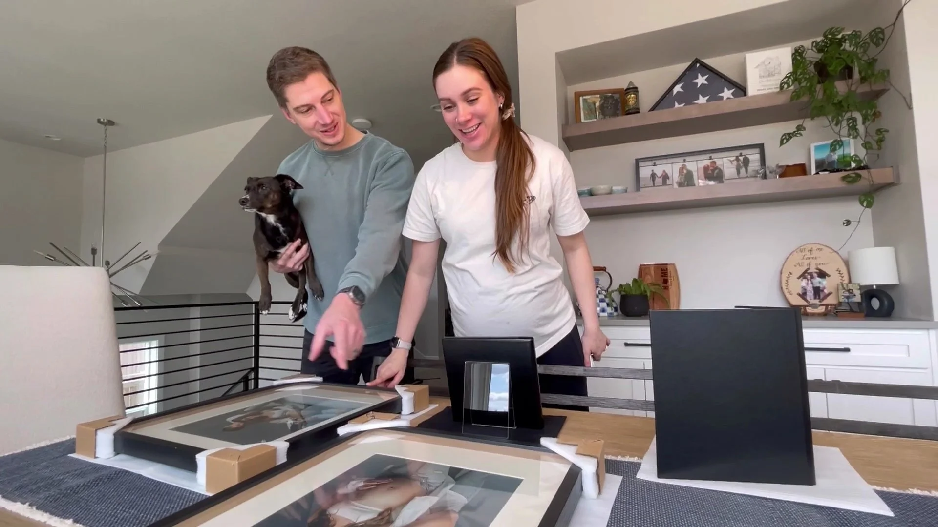A man holding a dog and a woman smiling while looking at newly delivered framed photos on a dining table.
