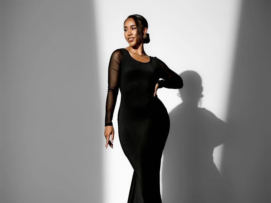 A woman in a form-fitting black dress with sheer sleeves posing confidently against a white studio background with dramatic light and shadows.