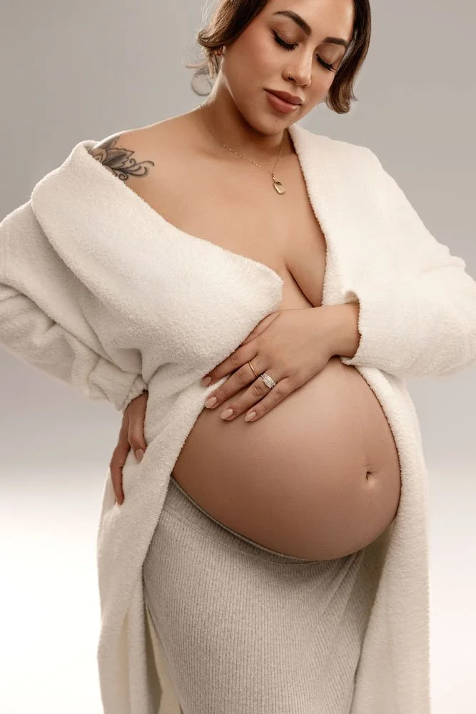 Sunnymelon Photography intimate fine art maternity portrait of an expectant mother in a cozy white robe and beige skirt, cradling her bare pregnant belly in a close-up studio shot.