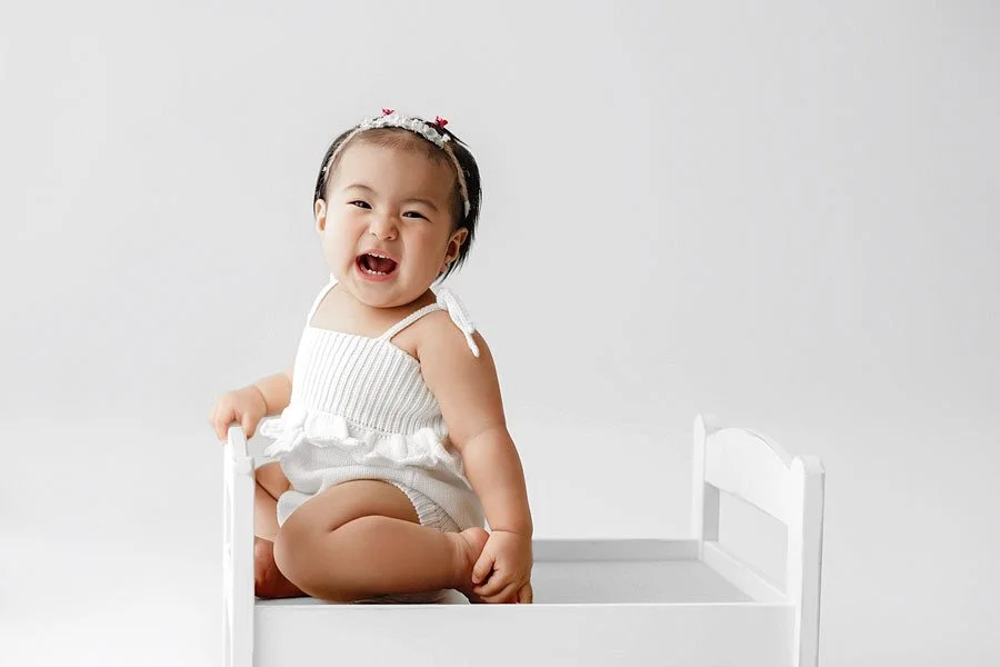 A laughing baby girl wearing a white knit romper and a floral headband sitting on a small white wooden bed in an all-white photography studio.