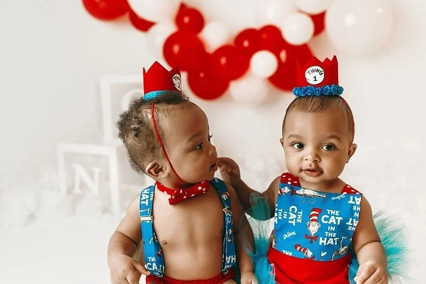 Twin babies in Dr. Seuss "Thing 1" and "Thing 2" themed outfits sitting together, with one baby looking at the other during their first birthday photoshoot.