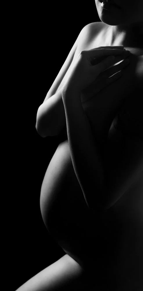 Sunnymelon Photography intimate black and white fine art maternity portrait. The expectant mother's pregnant belly and silhouette are dramatically lit from the side against a deep black background, emphasizing the curve of her body and hands.