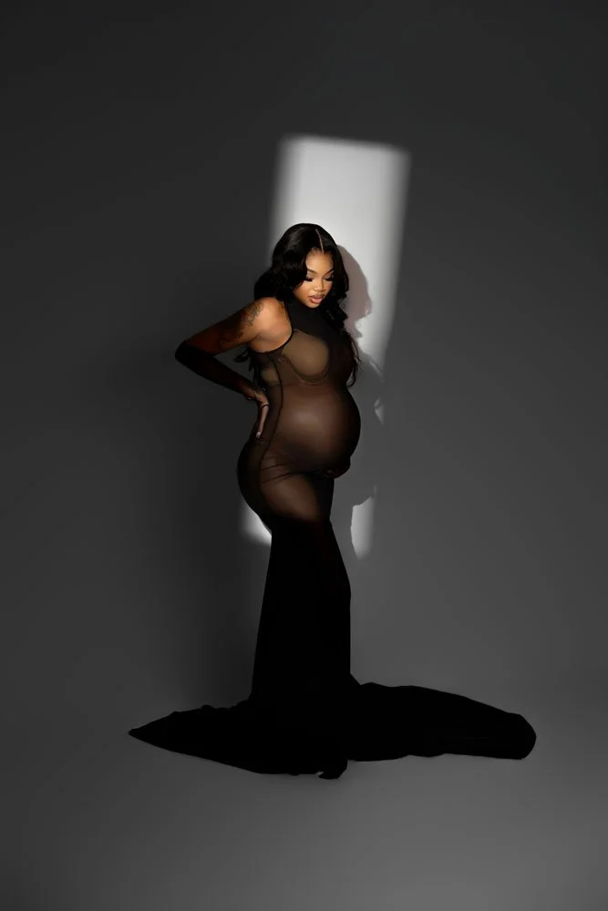 An editorial studio maternity portrait of a pregnant woman in a sheer black gown and opera gloves against a dark grey backdrop in West Linn, Oregon. A harsh vertical beam of spotlight illuminates her baby bump in a dramatic, high-contrast pose.