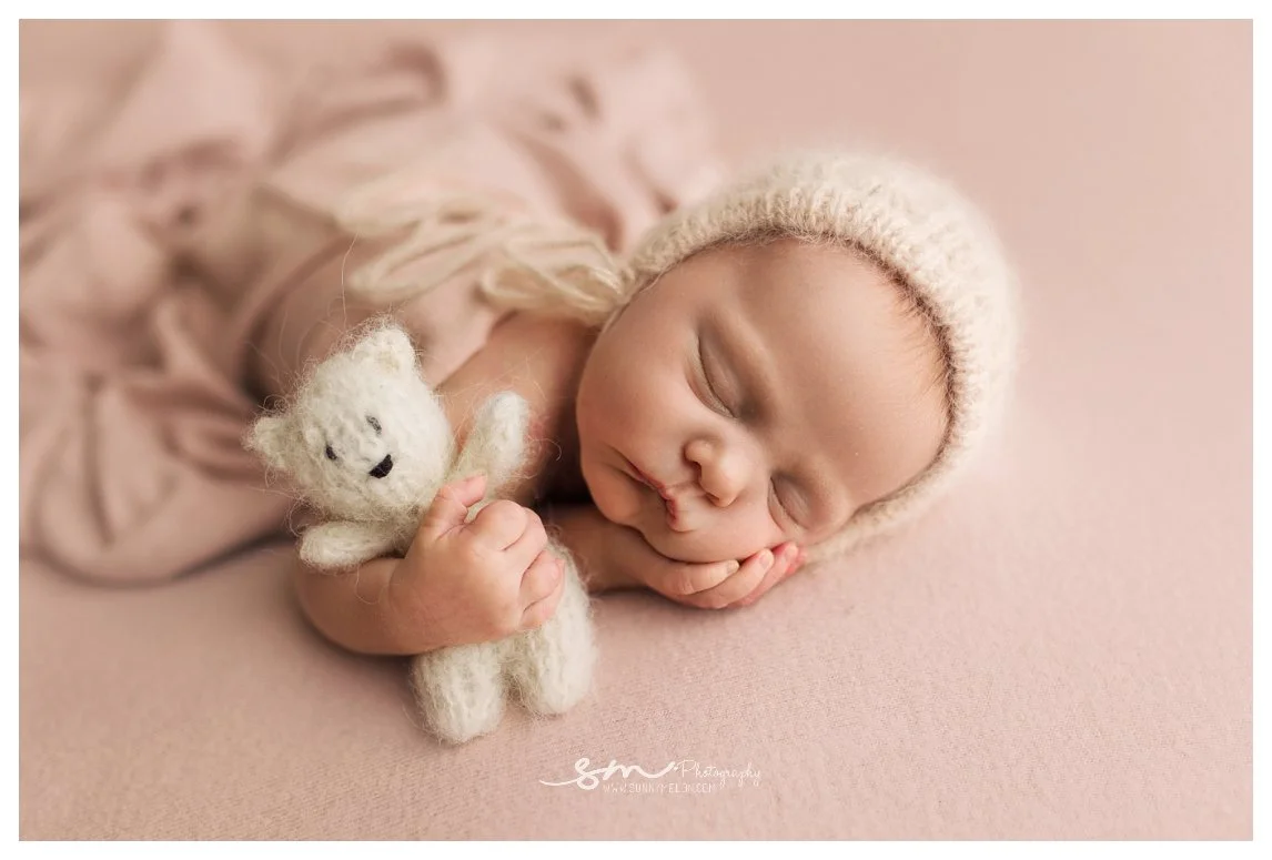 A close-up studio newborn portrait against a soft pink background showing a sleeping baby wearing a fuzzy knit bonnet and cradling a tiny white knit teddy bear.