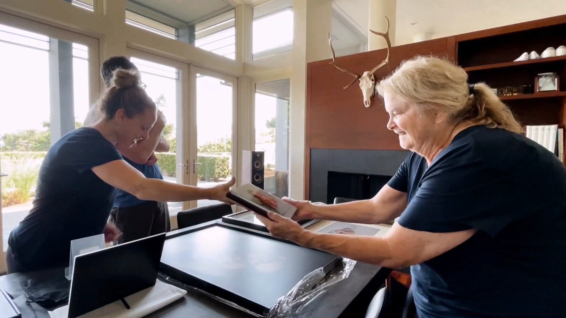A smiling woman in a dark t-shirt presents a high-quality, custom-made photo album to a client in a bright, modern living room. The image showcases the premium heirloom products and personalized gallery reveal service offered by a Portland family pho
