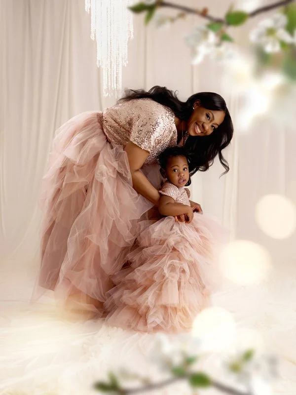 A mother and her young daughter posing in matching rose gold sequin tops and voluminous blush pink tulle skirts in a bright, ethereal studio setting with a crystal chandelier.