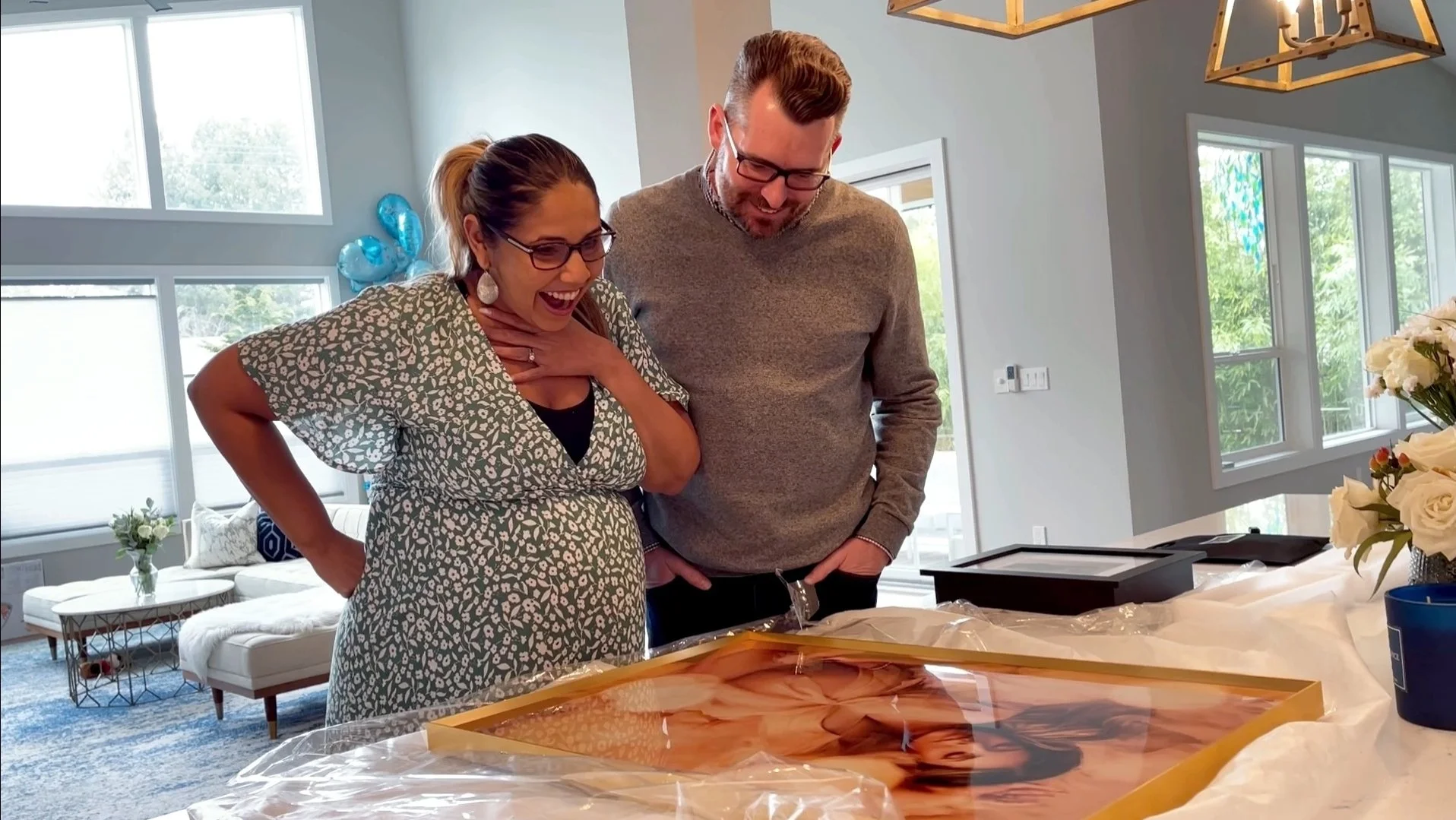 A candid moment of a surprised and happy couple unboxing a large, framed fine-art portrait in their home. The mother is smiling with her hand to her chest, showcasing the high-quality physical products and gallery reveal experience provided by a Port