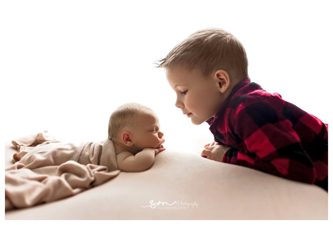A backlit studio newborn portrait against a bright white background showing a toddler boy in a red and black plaid shirt leaning in face-to-face with his sleeping newborn sibling who is wrapped in a neutral cloth.
