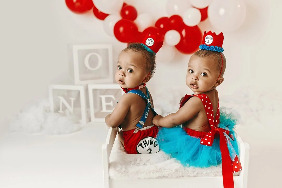 This adorable twin milestone portrait offers a charming "over-the-shoulder" view of the "Thing 1 and Thing 2" first birthday session. The twins are posed together on a miniature white bench, showcasing the back details of their custom Dr. Seuss outfi