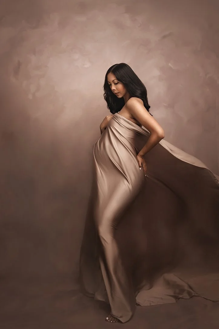 Elegant maternity photoshoot in Portland, Oregon, featuring a woman in a flowing silk dress, cradling her baby bump, with a soft brown background.