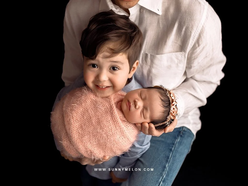 A toddler boy smiles warmly while holding his newborn baby sister, who is sleeping in a soft pink knit swaddle with a floral headband, against a dark studio background.