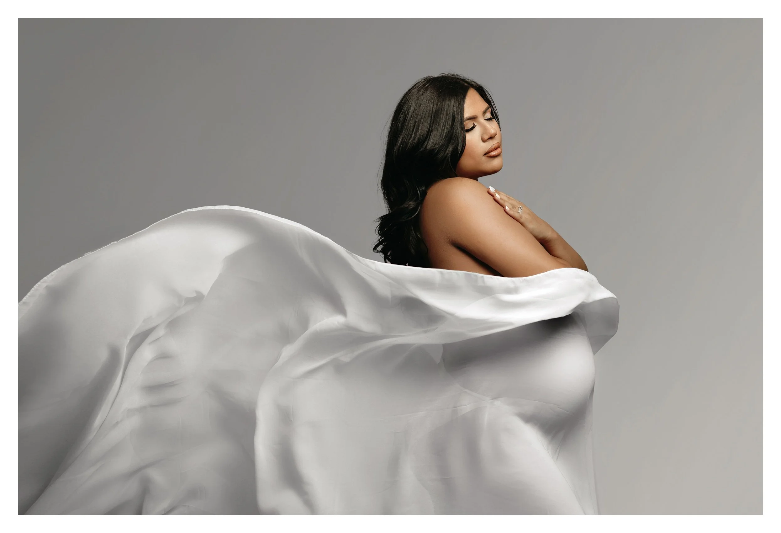 A fine art studio portrait against a grey backdrop showing a pregnant woman in profile with her eyes closed; translucent white fabric billows around her shoulders and drapes over her baby bump.