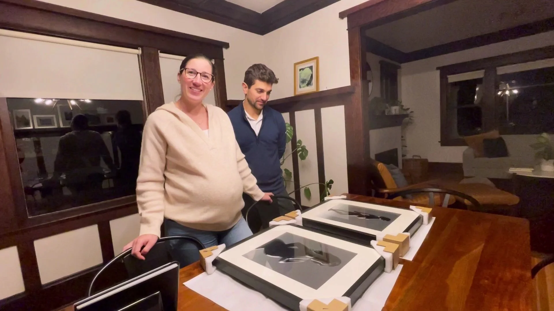 A pregnant woman and a man standing over a wooden dining table looking at two newly unboxed, framed photographs.