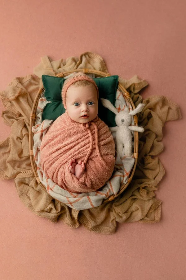 A professional overhead studio portrait by SunnyMelon Photography featuring a wide-eyed newborn baby girl. The infant is snugly wrapped in a textured rose-pink swaddle with a matching knit bonnet, resting in a small wicker basket. She is posed on a d